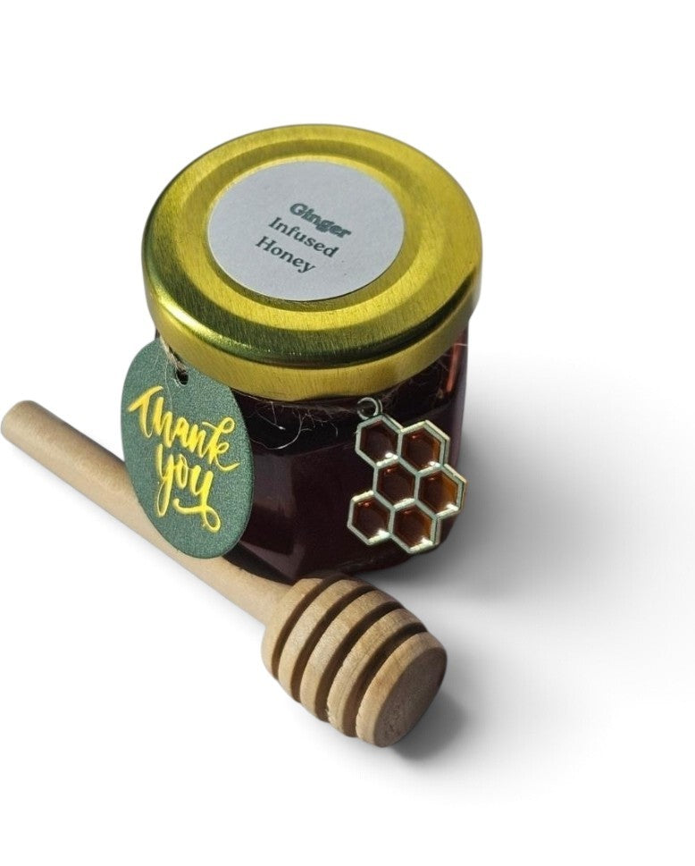 A small jar of ginger infused honey with a gold lid, a green 'thank you' tag, and a wooden honey dipper on a white background.