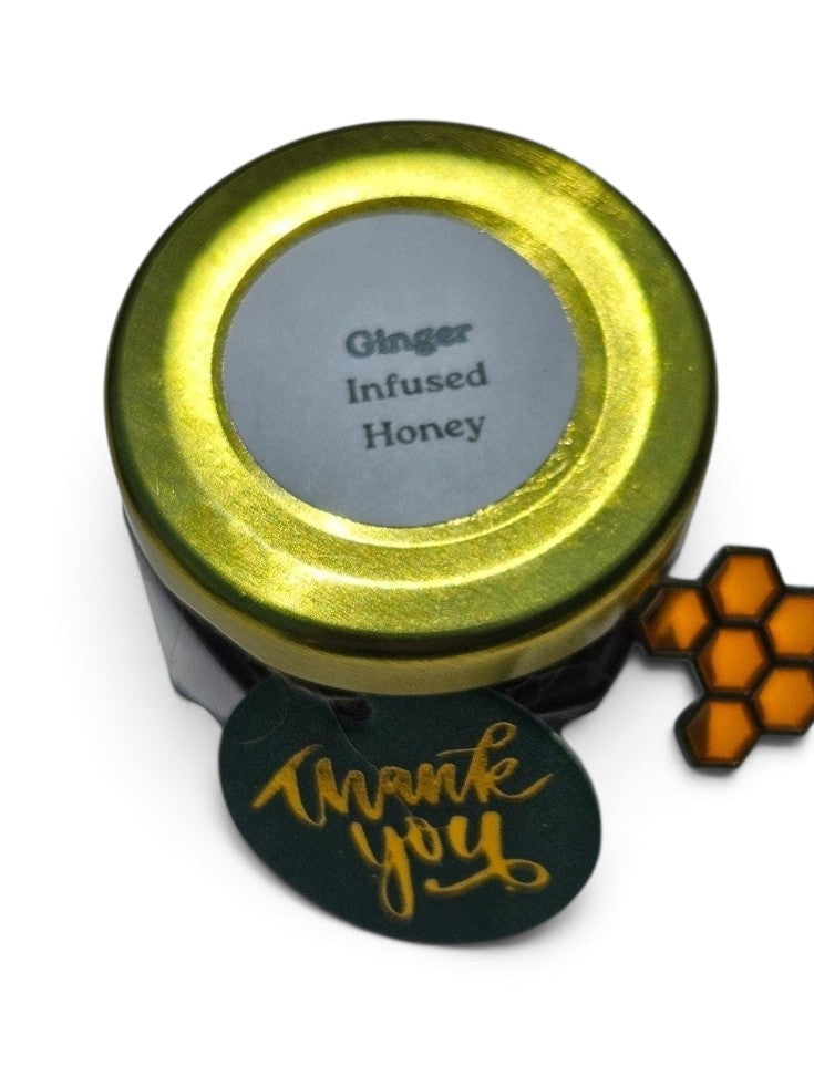 A small round jar of ginger infused honey with a metallic gold lid and a white label reading 'Ginger Infused Honey.' A dark green round tag with gold script says 'thank you,' and an orange honeycomb-shaped tag is attached on the right, all on a white background.