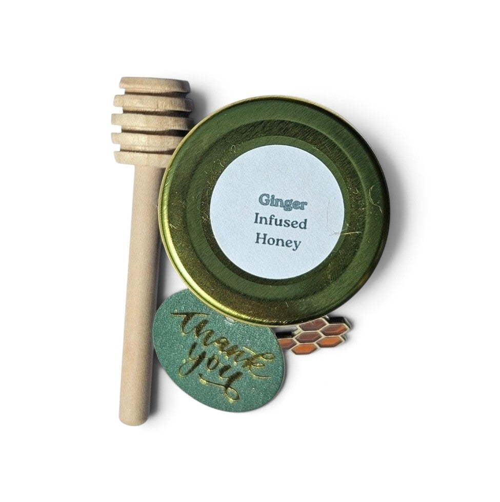 A ginger infused honey sample jar featuring a small, round, white label, a wooden honey dipper, and a round green tag with gold foil 'thank you' script.