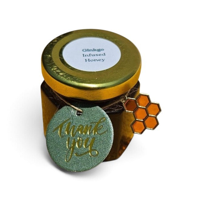 A small jar of Ginkgo Infused Honey with a gold lid and a round green glittery 'thank you' tag hanging from the side, alongside an orange honeycomb charm, on a white background.