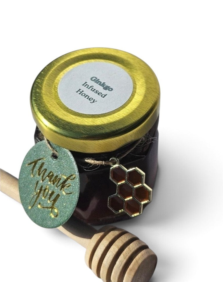 A jar of Glikage Infused Honey with a gold lid, a green 'thank you' tag, and a honeycomb charm, shown with a wooden honey dipper on a white background.