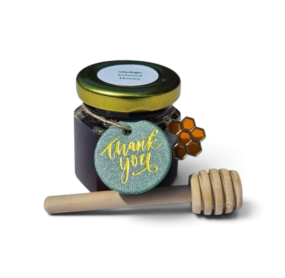 A small jar of Ginkgo honey with a gold lid, a wooden honey dipper, and a round glittery teal 'thank you' tag with a honeycomb charm, photographed on a white background.