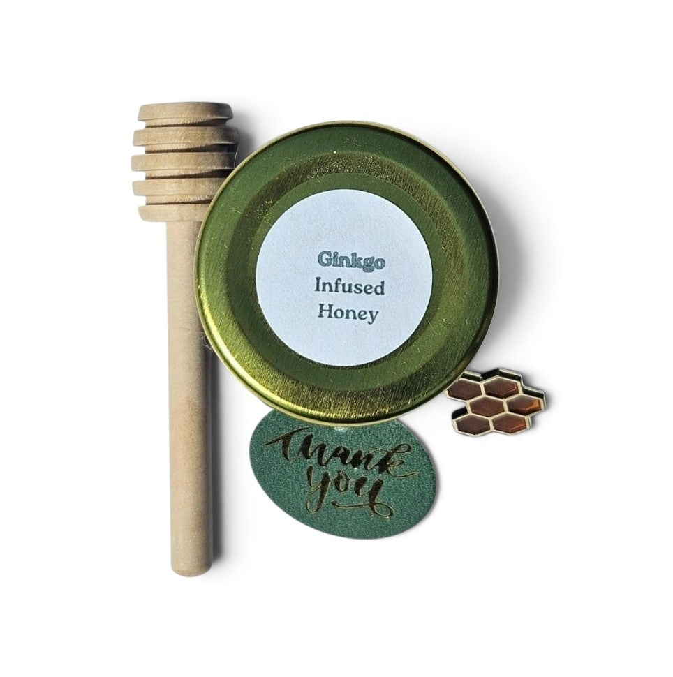 A sample jar of honey featuring a wooden honey dipper, a round gold lid with a white label that says Ginkgo Infused Honey, a green round thank you tag and an accessory shaped like a honeycomb.