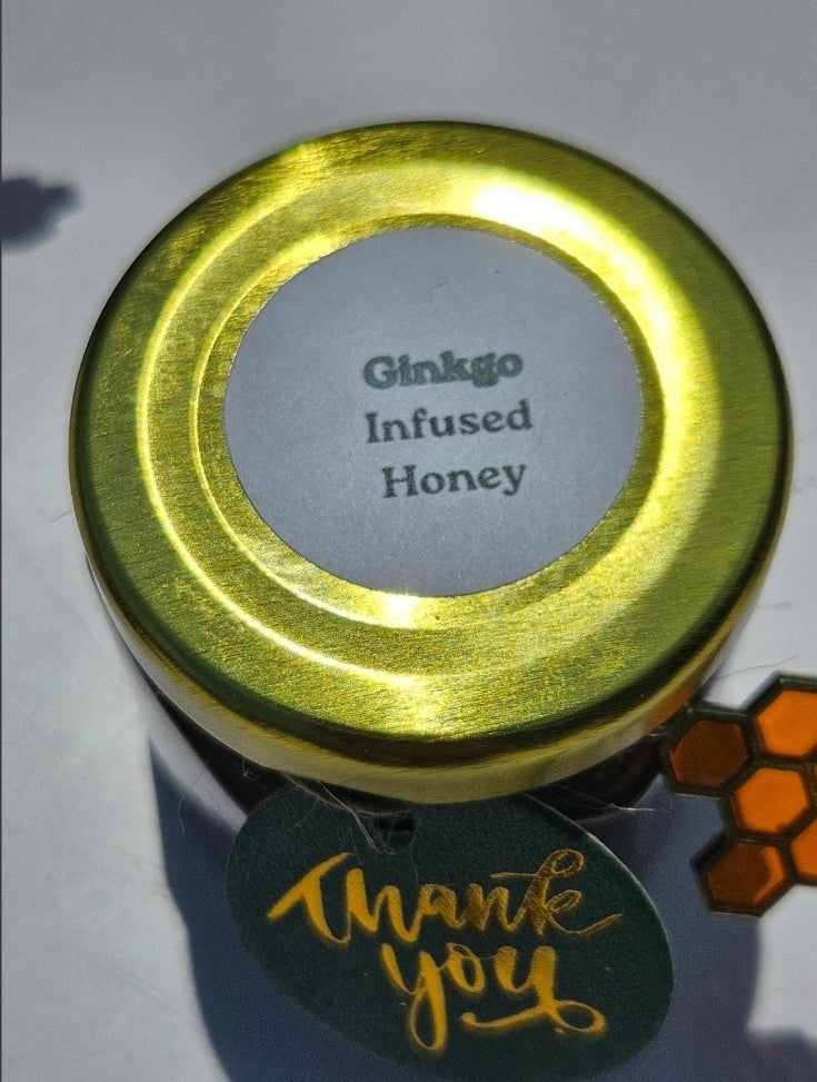 A small jar of Ginkgo Infused Honey with a shiny gold lid and a gray label. A dark green 'thank you' tag with gold script is attached to the jar, and a small orange honeycomb-shaped tag is visible on the right side, all on a light gray background.