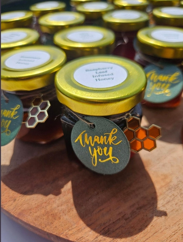 Small jars of honey with gold lids arranged on a wooden surface, each decorated with a round green 'thank you' tag and a small honeycomb charm.