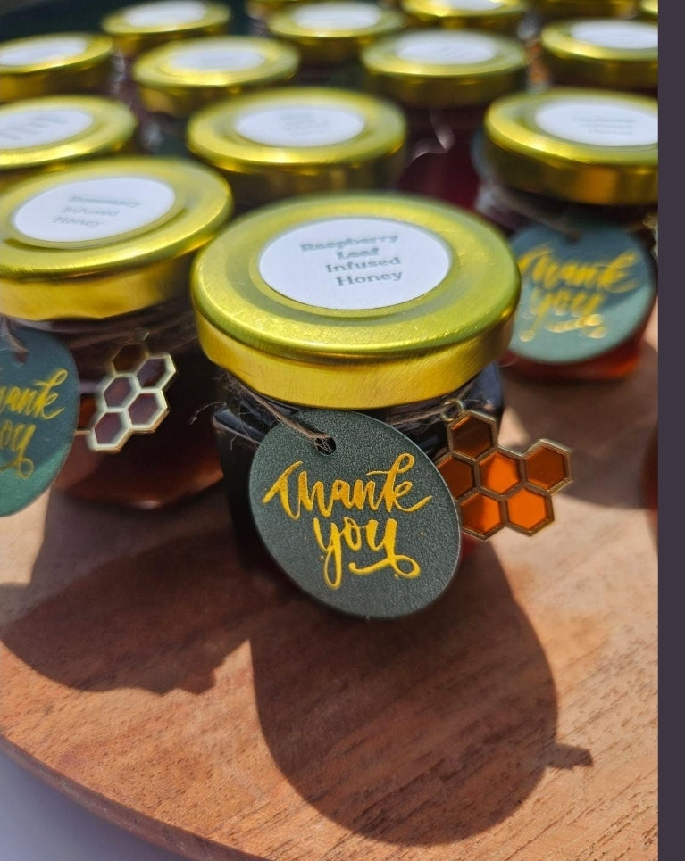 A close-up of several small glass jars of honey with gold lids arranged on a wooden surface. The front jar has a round dark green tag that says "thank you" in gold script, with a small honeycomb charm hanging beside it.