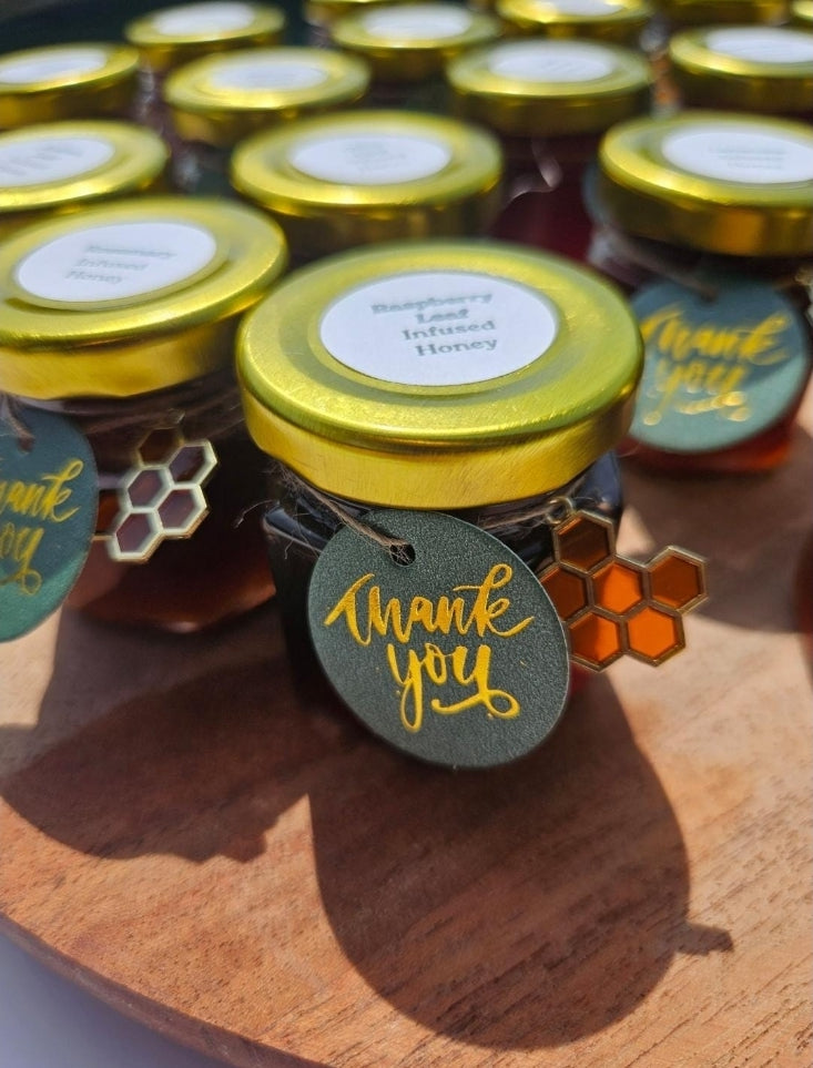 Several small glass jars of honey with gold lids are arranged on a wooden surface. The front jar has a round white label and a dark green hanging tag that says thank you in gold script, with a small orange honeycomb charm.