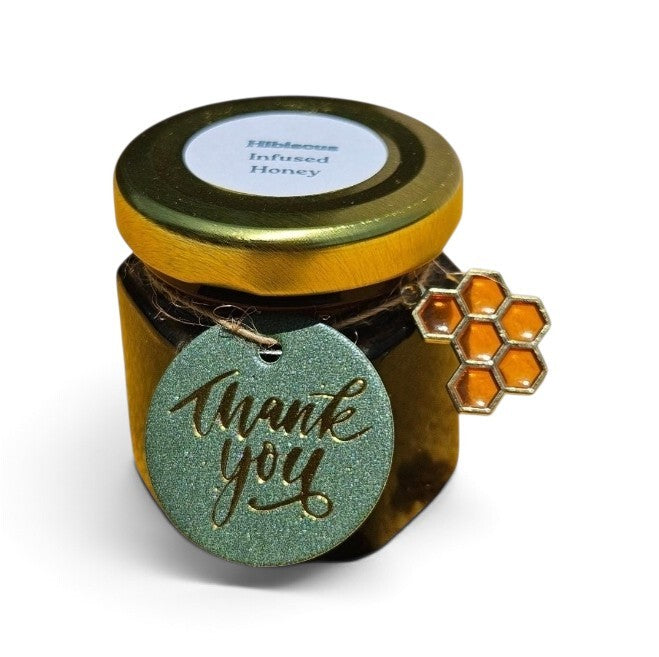 A small jar of hibiscus infused honey with a gold lid and a round green 'thank you' tag tied to the side, plus a small honeycomb charm, on a white background.