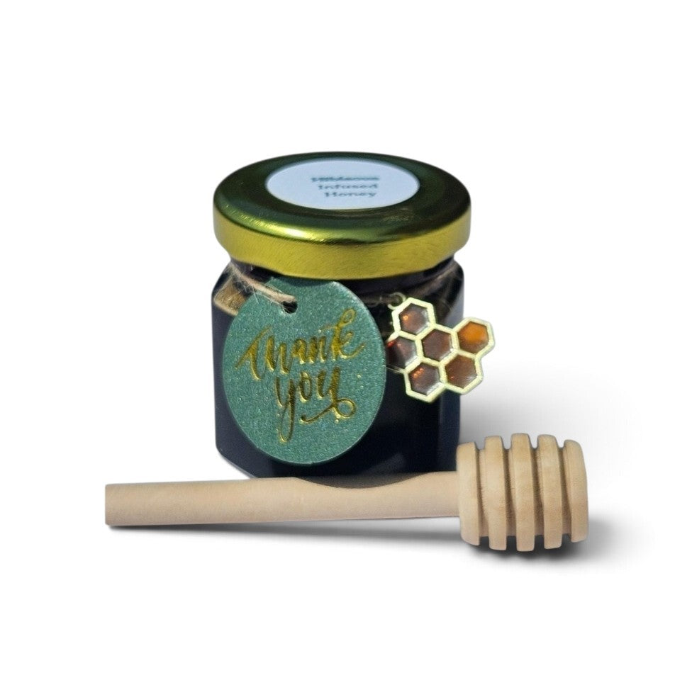 A small jar of hibiscus infused honey with a gold lid, a green glittery 'thank you' tag, a honeycomb charm, and a wooden honey dipper on a white background.