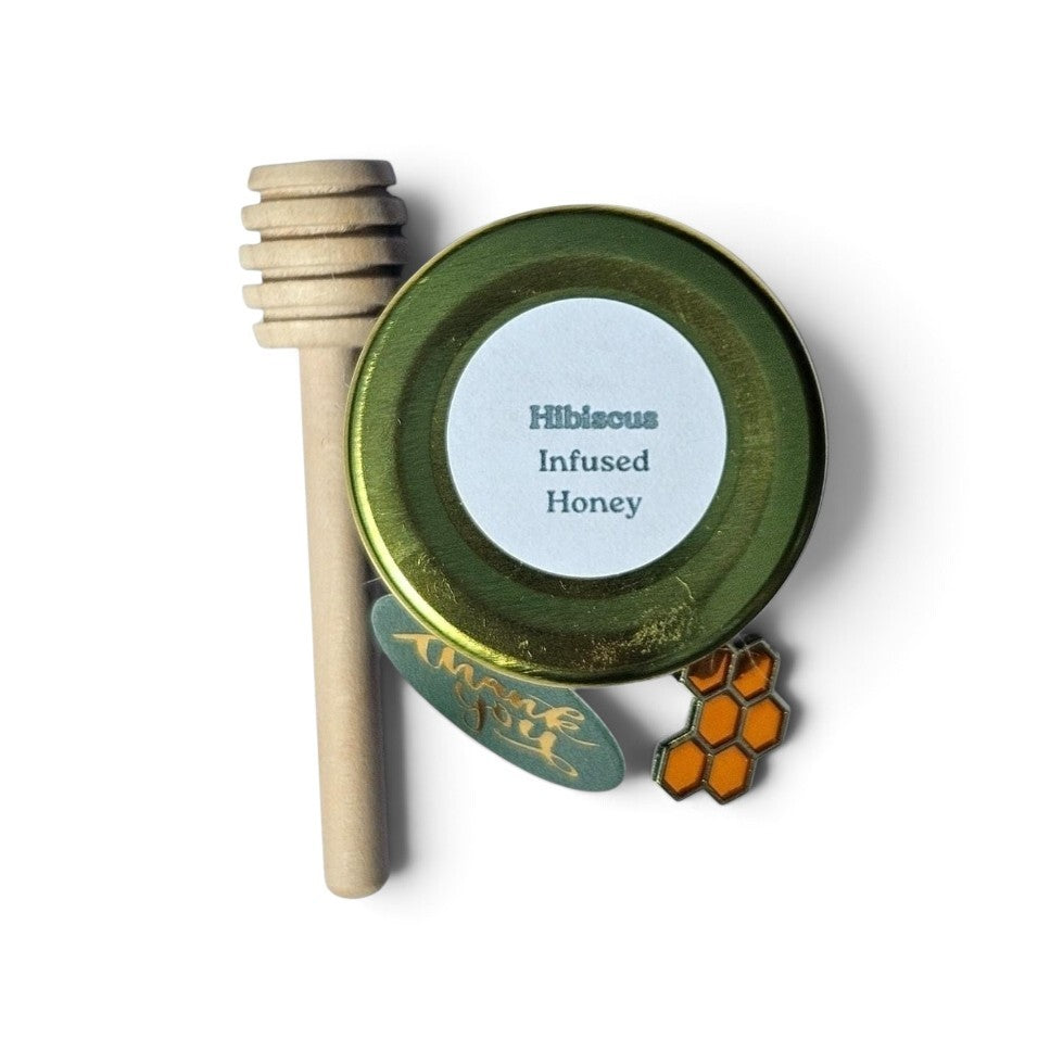 A small round jar of Hibiscus Infused Honey with a gold lid, shown next to a wooden honey dipper and honeycomb accessory. 