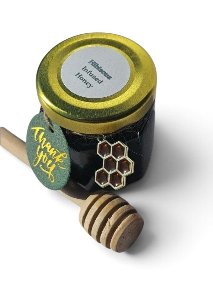 A sample jar of Hibiscus Infused Honey with a gold lid, a green 'thank you' tag, and a wooden honey dipper on a white background.