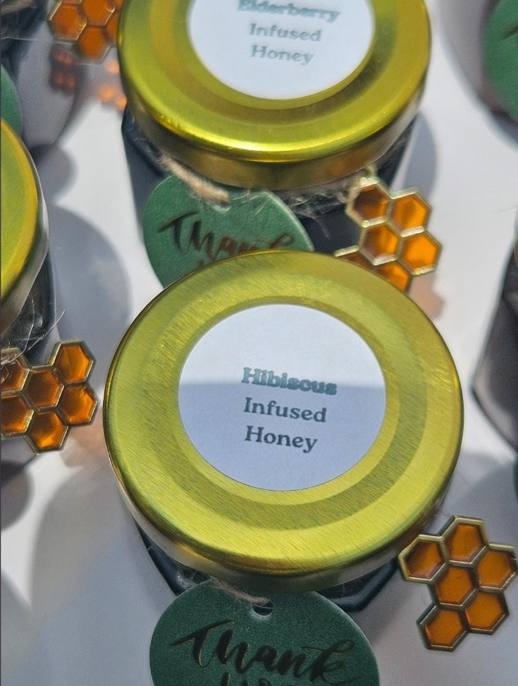 Two glass jars of infused honey with shiny gold lids, one labeled 'Elderberry Infused Honey' and the other 'Hibiscus Infused Honey', decorated with green tags and orange honeycomb charms on a white background.