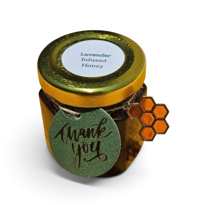 An amber glass jar of lavender infused honey with a shiny gold lid, featuring a green glittery 'thank you' tag and an orange honeycomb sticker on the side.