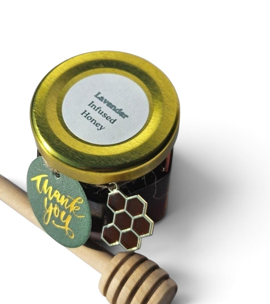A small jar of lavender infused honey with a gold metal lid, a round white label, a green 'thank you' tag, and a honeycomb-shaped charm, shown with a wooden honey dipper on a white background.