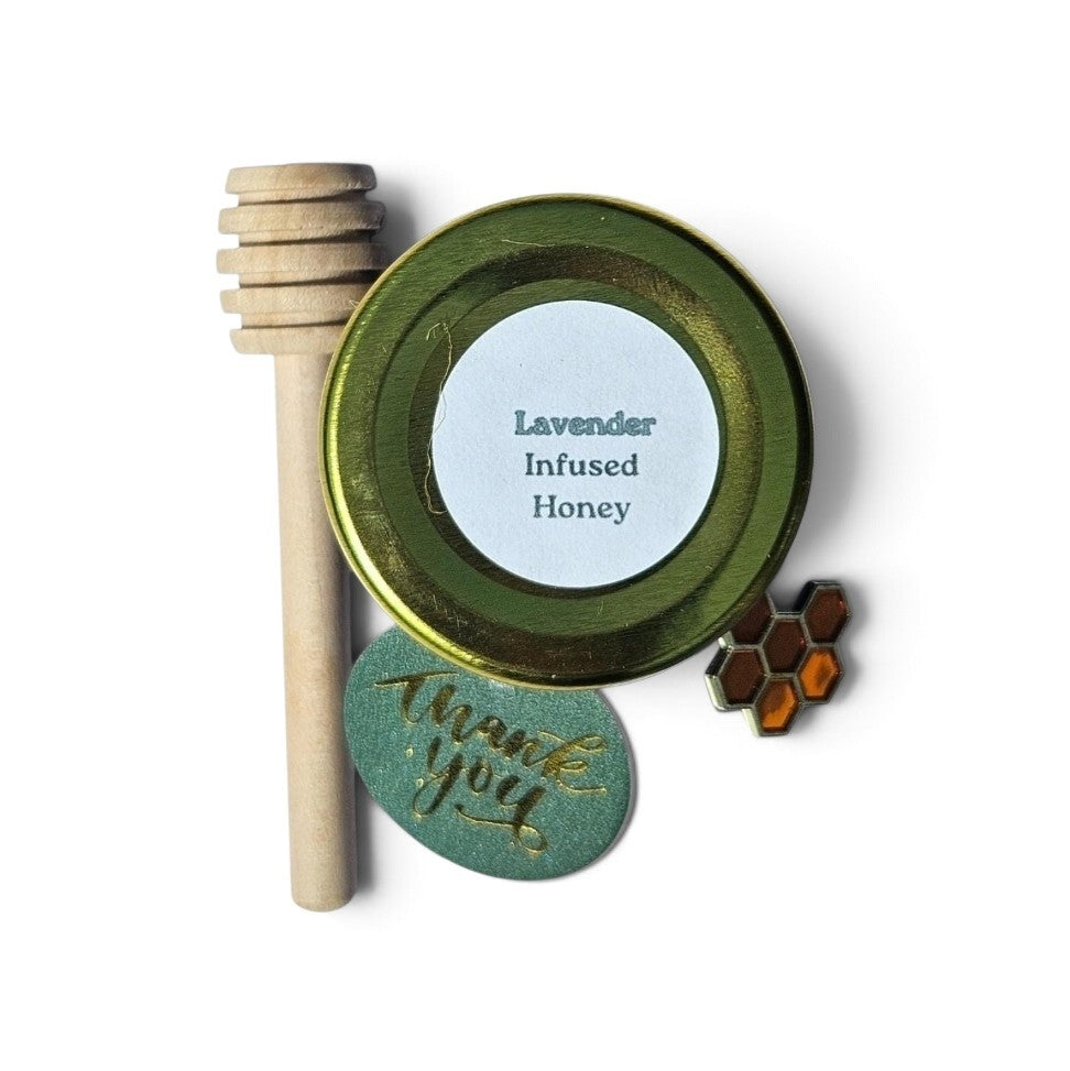 A gift set of infused honey featuring a round gold lid  labeled 'Lavender Infused Honey,' a wooden honey dipper, a round green tag with gold 'thank you' script, and a small honeycomb charm, arranged on a white background.