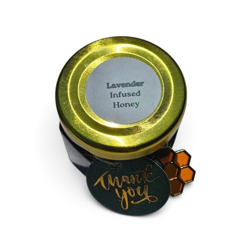 A small jar of lavender infused honey with a shiny gold lid and a green tag that reads 'thank you' in gold script, alongside a small honeycomb charm, on a white background.
