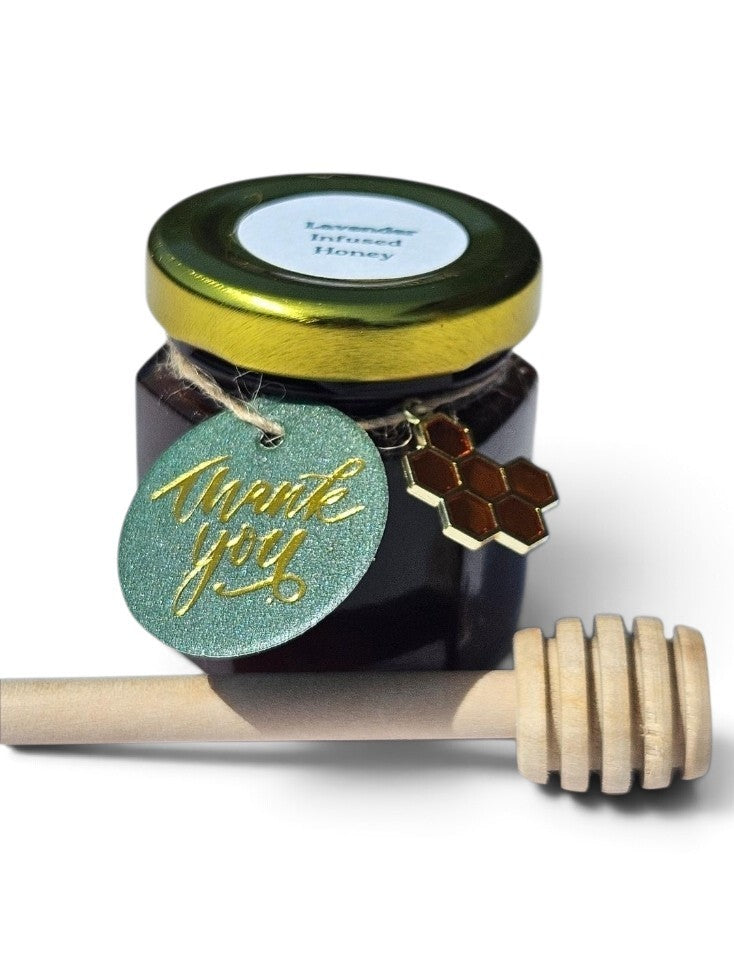 A jar of lavender infused honey with a shiny gold lid, decorated with a glittery green 'Thank You' tag and a honeycomb charm, with a wooden honey dipper resting in front on a white background.
