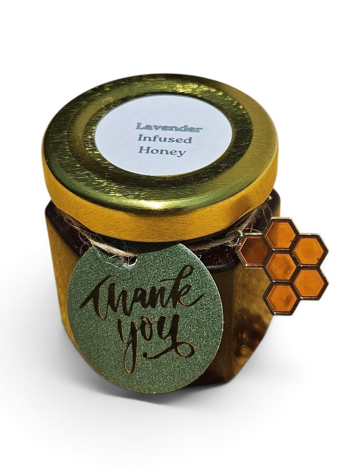 A small glass jar of lavender infused honey with a shiny gold lid and a green glittery 'thank you' tag on the front, alongside an orange honeycomb-shaped accent, set against a plain white background.