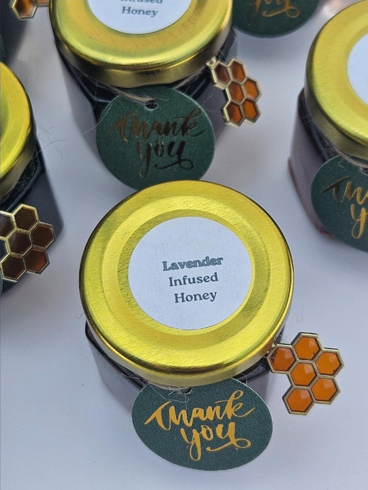 Several small jars of honey with shiny gold lids are arranged on a white surface. The center jar is labeled 'Lavender Infused Honey' and has a dark green round tag reading 'thank you' with gold script, plus small honeycomb-shaped embellishments.