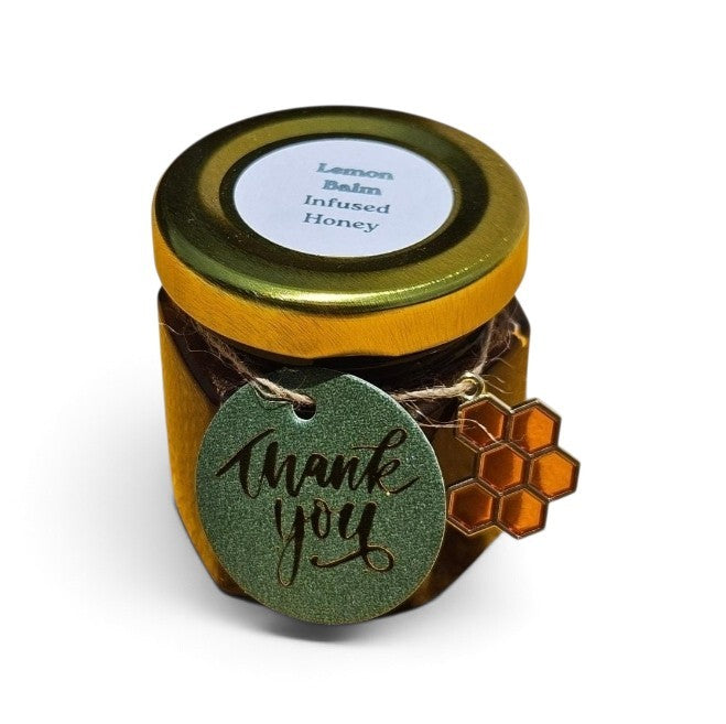 A small glass jar of lemon balm infused honey with a gold lid, decorated with a green 'thank you' tag and a honeycomb charm, on a white background.