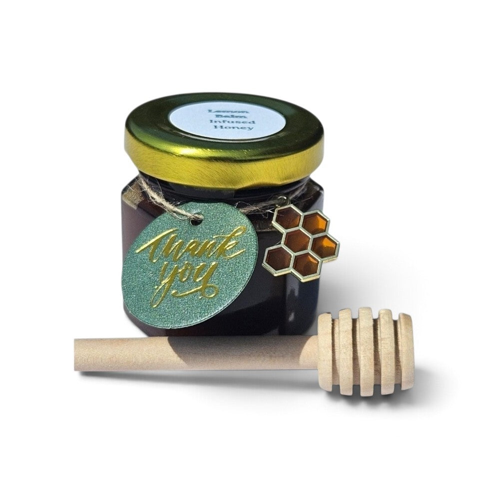 A jar of honey with a gold lid, a green glittery 'thank you' tag, and a wooden honey dipper on a white background.