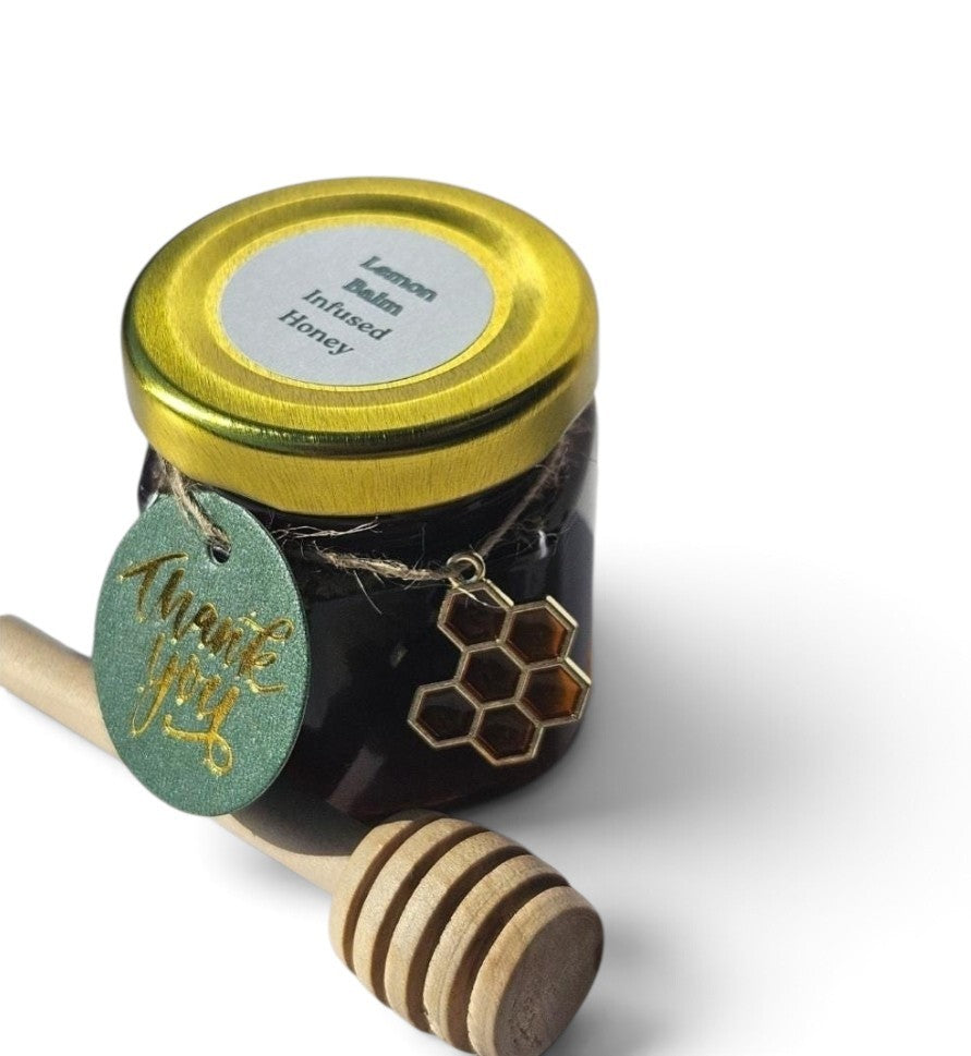 A small glass jar of honey with a gold lid and a round green hang tag reading 'thank you,' shown with a wooden honey dipper on a white background. The jar label reads 'Lemon Balm Infused Honey.'