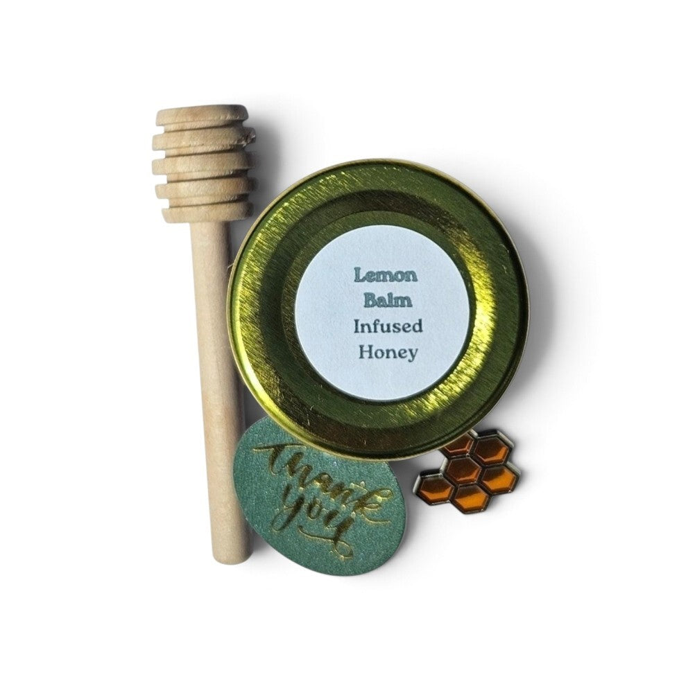 A sample sized jar featuring a small round label of 'Lemon Balm Infused Honey' with a white label, a wooden honey dipper, a green 'thank you' card, and a small honeycomb-shaped accessory, all arranged on a white background.