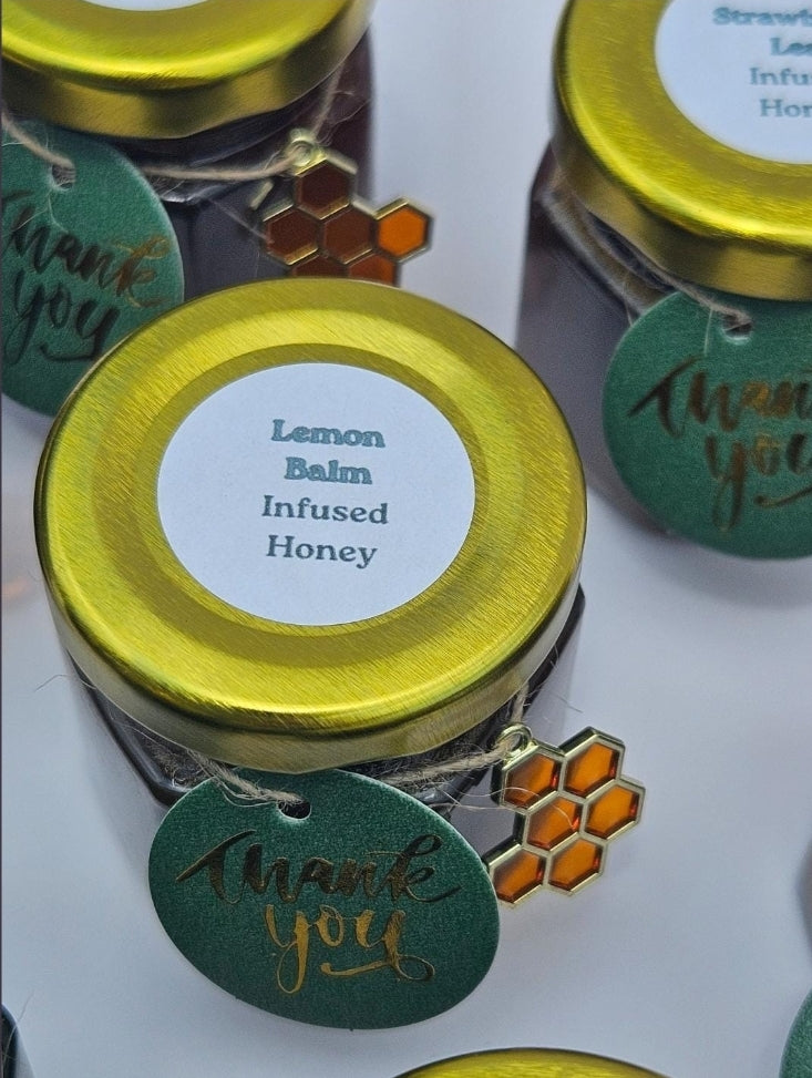 Small glass jars of infused honey with shiny gold lids, round white labels reading 'Lemon Balm Infused Honey,' and green oval hang tags that say 'thank you' with gold script. One tag also has a small orange honeycomb charm attached.