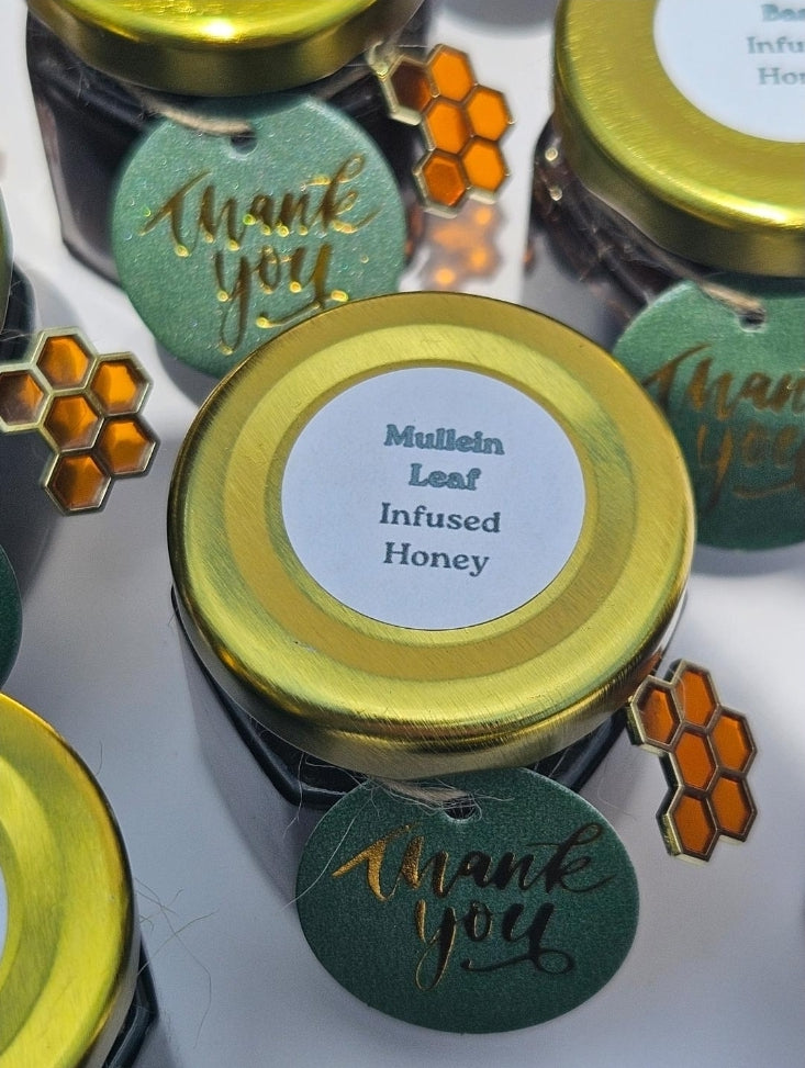 Several small jars of infused honey with gold lids are arranged on a white surface. The center jar has a white label reading 'Mullein Leaf Infused Honey' and a green hanging tag that says 'thank you.