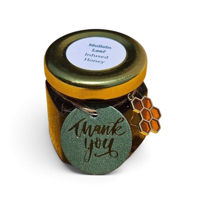 A small glass jar of mullein leaf infused honey with a gold lid and a round green 'Thank you' tag attached, plus a honeycomb charm, on a white background.