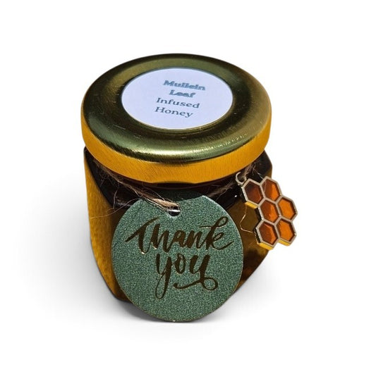 A small glass jar of mullein leaf infused honey with a gold lid and a round green 'Thank you' tag attached, plus a honeycomb charm, on a white background.