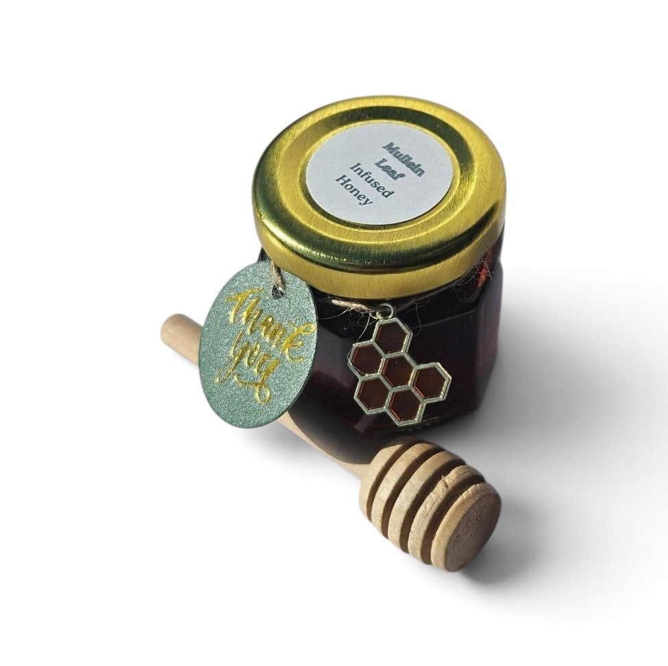A small glass jar of honey with a gold lid, a round white label reading 'Mullein Leaf Infused Honey,' a green 'thank you' tag, and a wooden honey dipper placed in front on a white background.