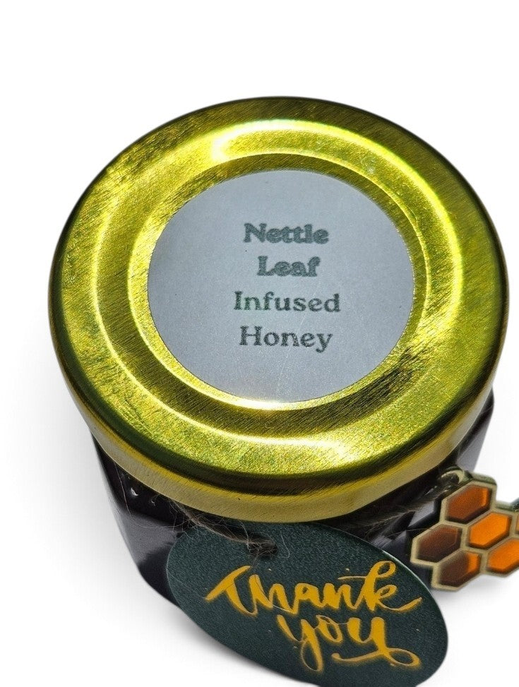 A jar of nettle leaf infused honey with a shiny gold lid and a dark glass body, shown on a white background with a hanging 'thank you' tag.