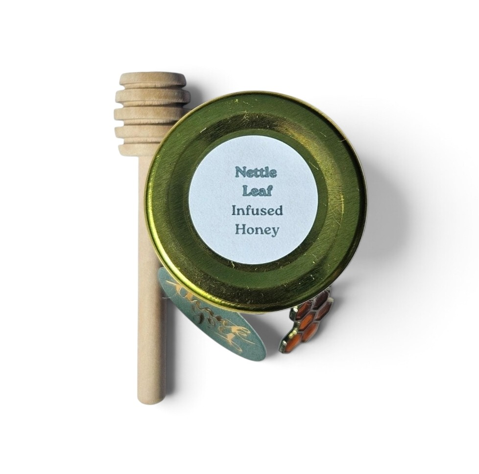 A round jar  of infused honey with a white label reading 'Nettle Leaf Infused Honey,' shown with a wooden honey dipper and a small honeycomb charm on a white background.