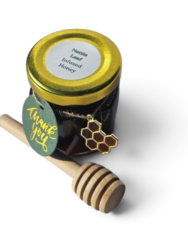 A small glass jar of infused honey with a shiny gold lid and a round green 'thank you' tag, shown with a wooden honey dipper on a white background. The jar label reads 'Nettle Leaf Infused Honey'.