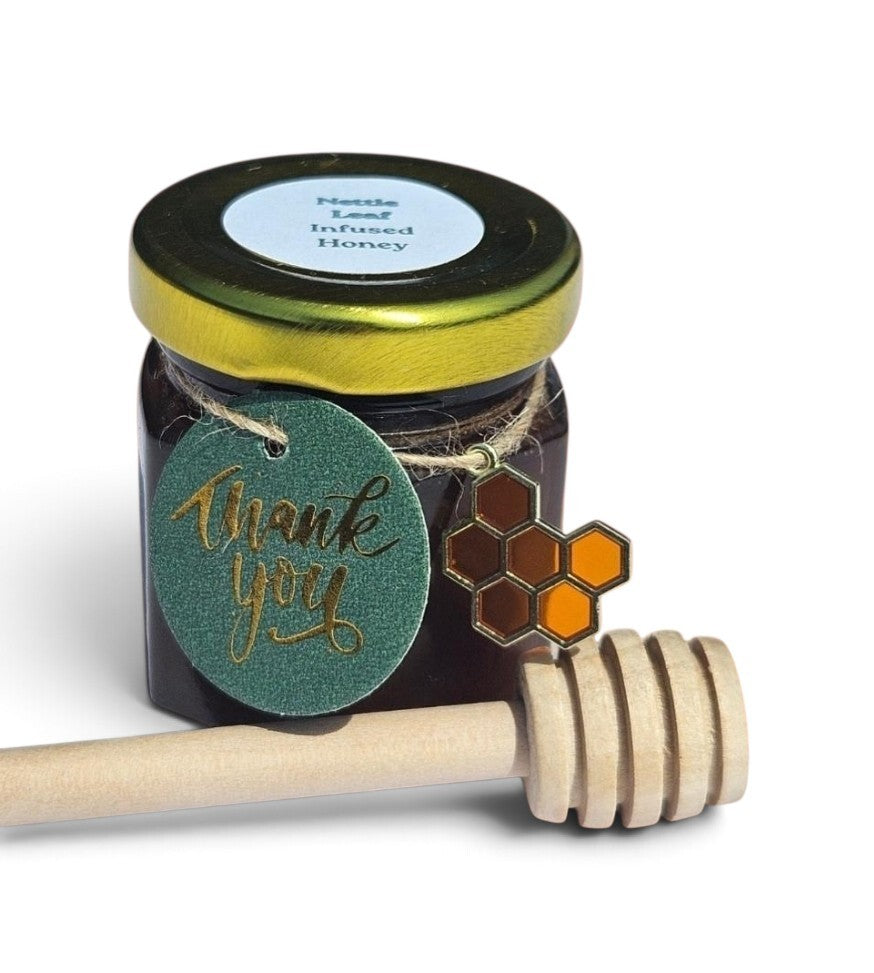 A sample jar of nettle leaf infused honey with a gold lid, a round green 'thank you' tag, a honeycomb charm, and a wooden honey dipper on a white background.