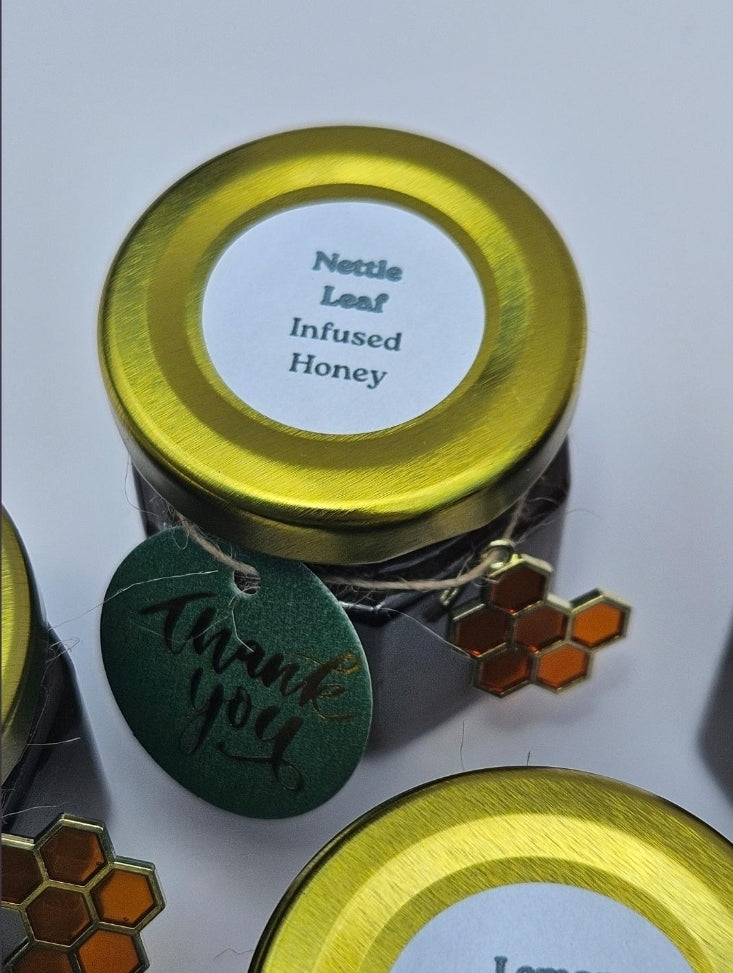A small jar of infused honey with a shiny gold lid and a white label reading 'Nettle Leaf Infused Honey,' shown on a light background with decorative tags and honeycomb charms.