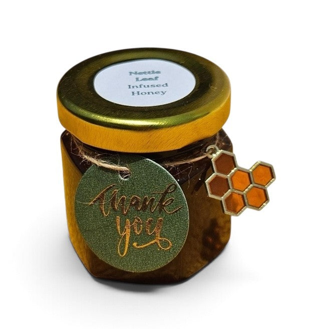 A small hexagonal glass jar of nettle leaf infused honey with a gold metal lid and a round green 'thank you' tag tied around the neck, featuring a honeycomb charm, on a white background.