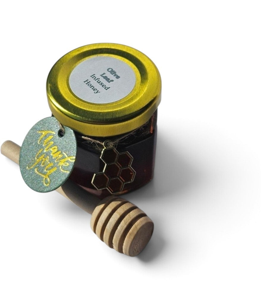 A small glass jar of honey with a gold metal lid and a white label reading 'Olive Leaf Infused Honey,' shown with a wooden honey dipper and a round green gift tag on a white background.