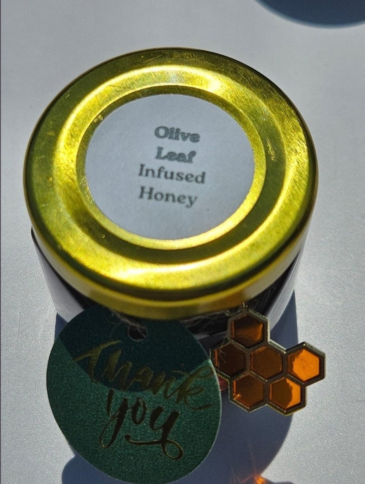 A small jar of infused honey with a shiny gold lid and a white label reading 'Olive Leaf Infused Honey.' A green round tag with 'thank you' in script and an orange honeycomb charm hang from the jar on a white background.