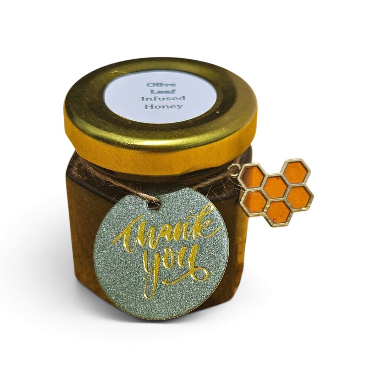 A small jar of olive leaf infused honey with a gold lid and a round glittery green 'thank you' tag hanging from the side, featuring an orange honeycomb charm, on a white background.