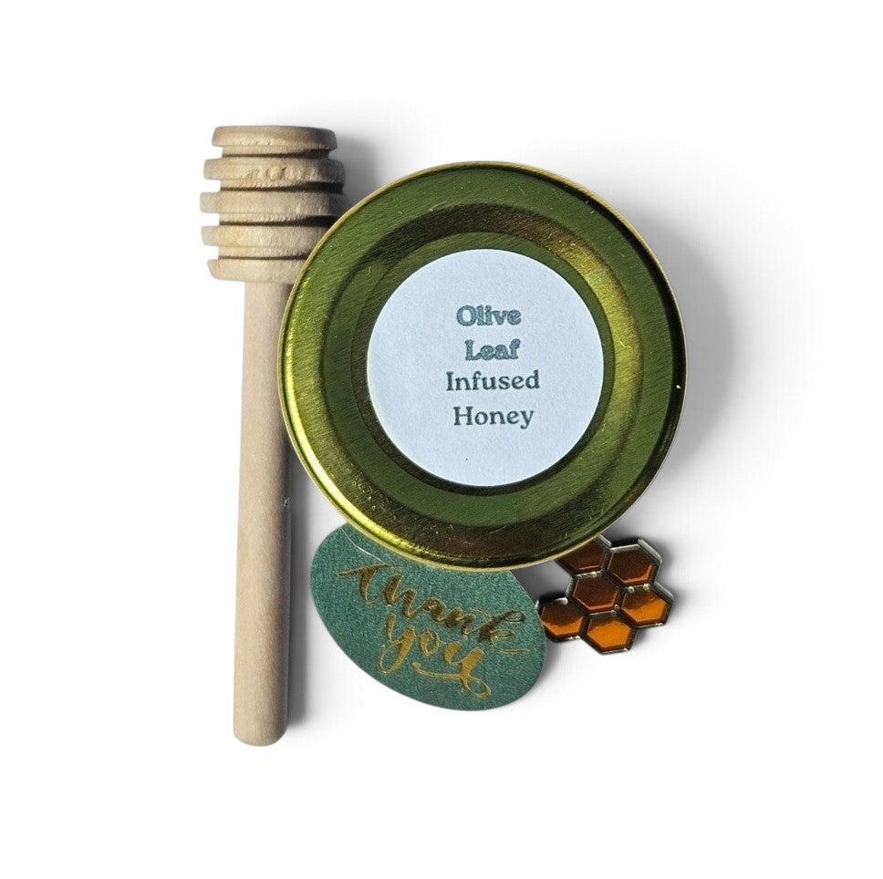 A sample sized jar of olive leaf infused honey with a wooden honey dipper and a small honeycomb-shaped accessory on a white background.