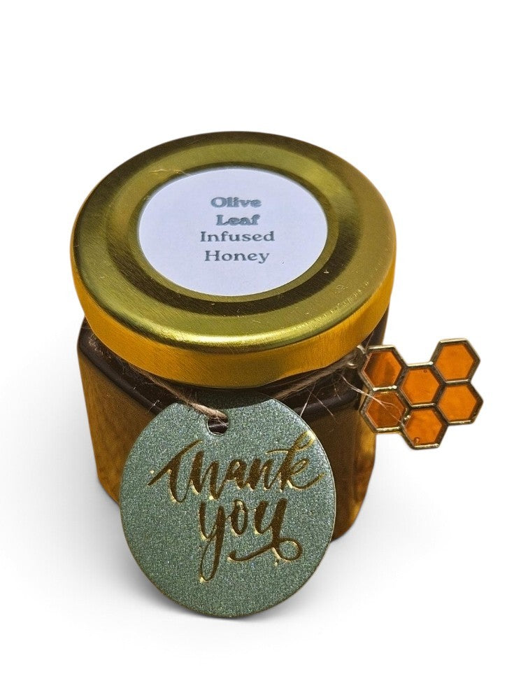 A jar of Olive Leaf Infused Honey with a gold metal lid, a white label, and a green glittery round tag that says 'thank you' with gold foil script. A small gold honeycomb charm is attached to the side.