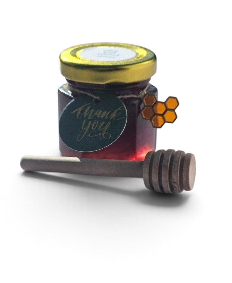 A small glass jar of dark, Olive Leaf infused honey with a gold lid and a round dark label reading 'thank you', shown with a wooden honey dipper in front on a white background.