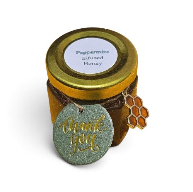 A small jar of Peppermint Infused Honey with a gold lid and a round green tag reading 'thank you' in gold script and a honeycomb charm, on a white background.