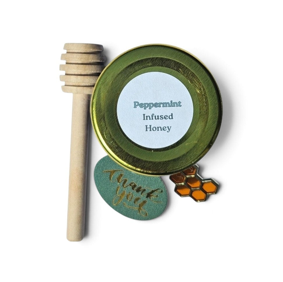 A sample jar of peppermint infused honey with a wooden honey dipper, a green 'thank you' tag, and a honeycomb-shaped charm on a white background.