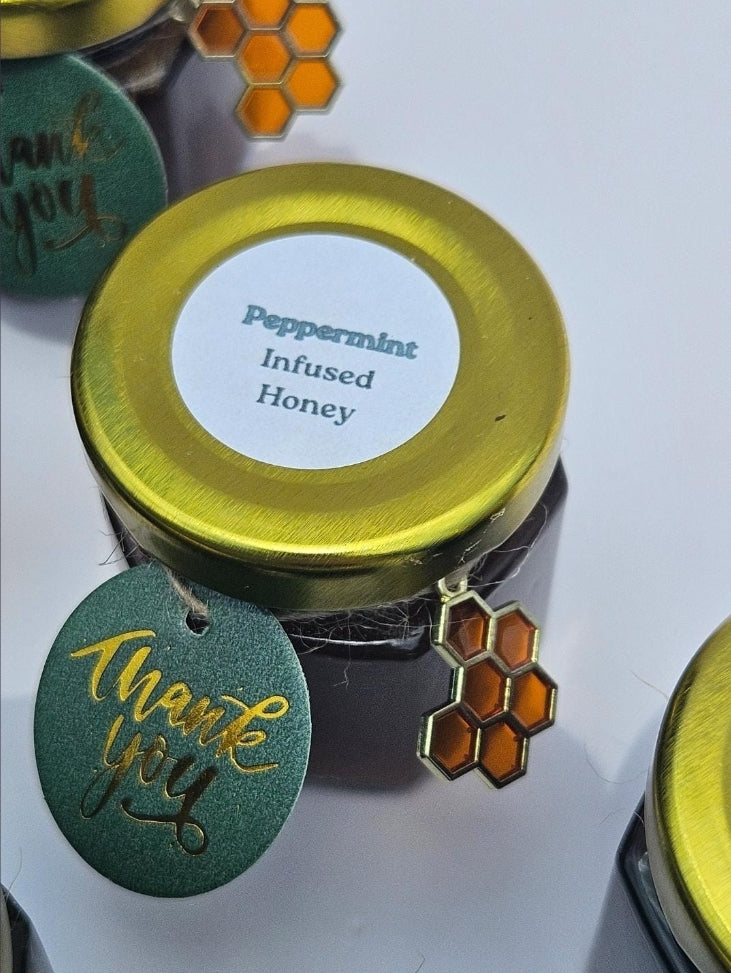 A small jar of infused honey with a shiny gold lid and a white label reading 'Peppermint Infused Honey.' A green round tag with gold script says 'thank you,' and a honeycomb charm hangs from the side.