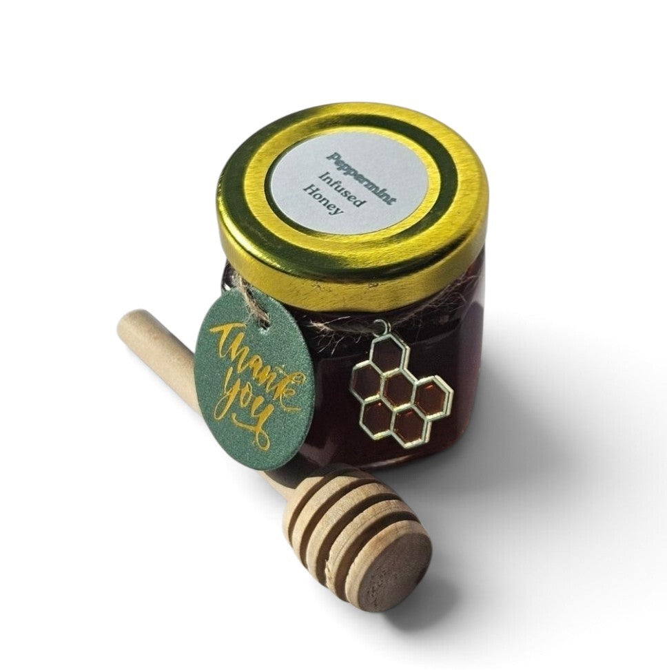 A small glass jar of Peppermint infused honey with a gold lid, a wooden honey dipper, and a green round tag that says 'thank you' in gold script, photographed on a white background.
