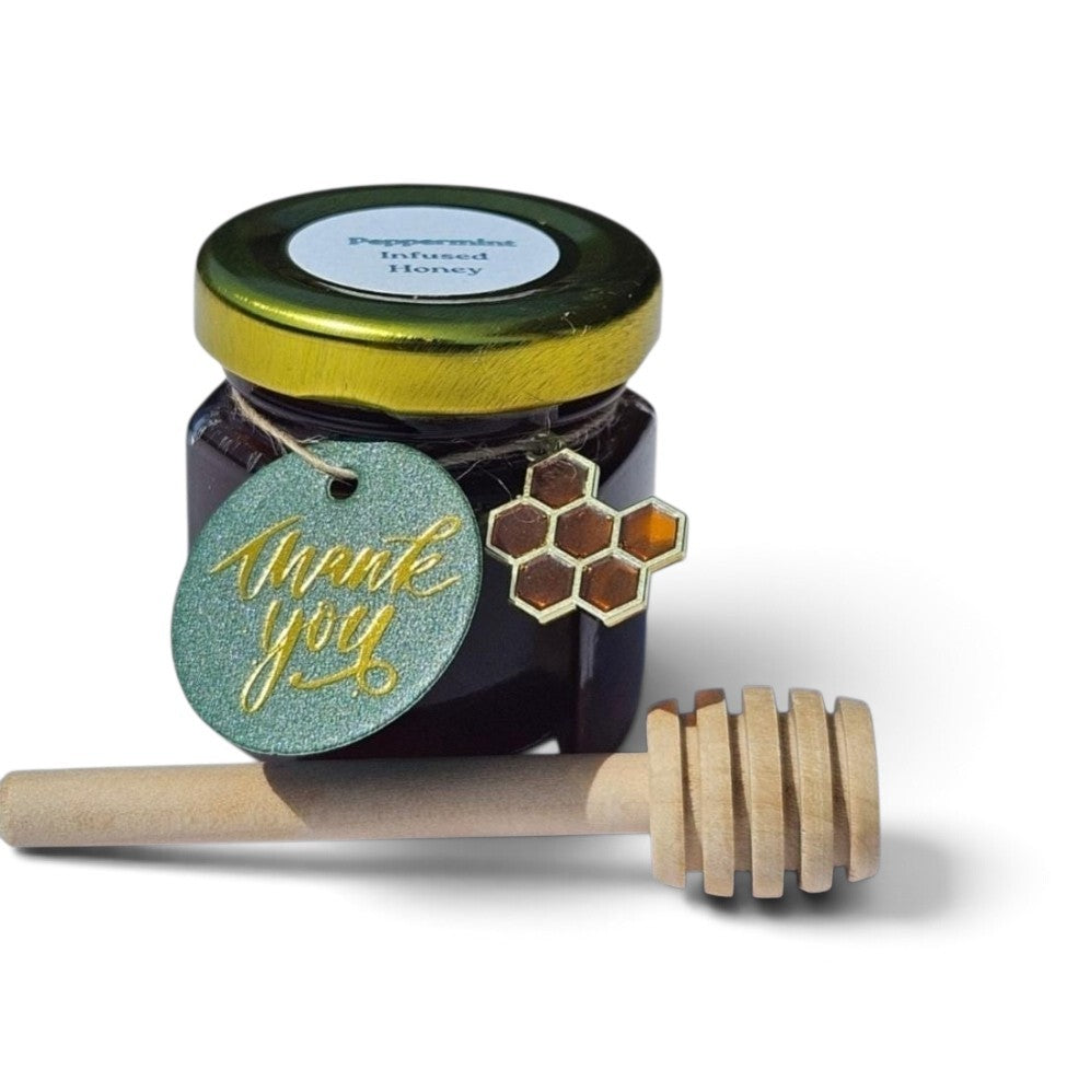 A jar of peppermint leaf infused honey with a gold lid, a round teal 'thank you' tag with gold script, a honeycomb-shaped charm, and a wooden honey dipper on a white background.