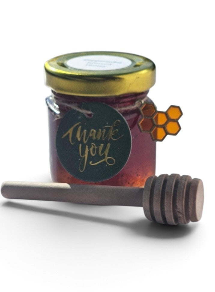 A small glass jar of honey with a gold metal lid and a round black 'Thank you' label, shown with a wooden honey dipper on a white background.